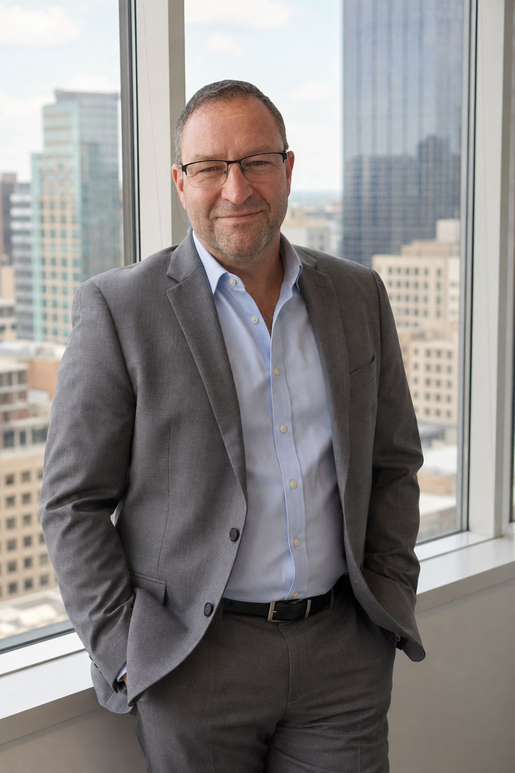Forrest Andrew Jones standing beside a high-rise office window in a grey jacket over an open-collar light blue shirt, with an out-of-focus city skyline behind the glass.
